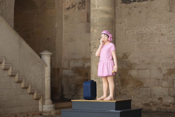 Adieu mes chers parents Enfant en robe rose, debout sur un socle, tenant une valise, dans un espace intérieur.